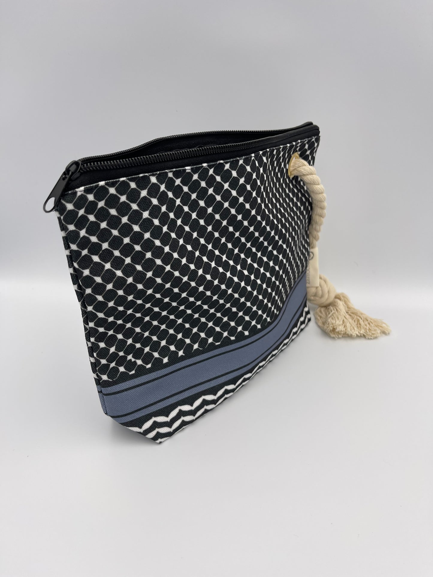 Angle shot of black makeup bag with beige rope against a white background. Bag is patterned with black keffiyeh design and ZEITOUNI logo tag.