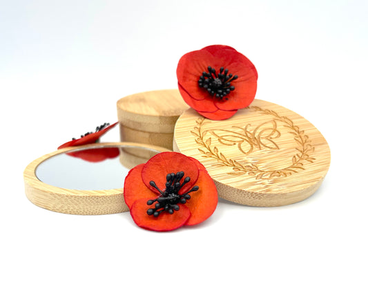 Bamboo mirrors stacked with red poppy flowers against a white background. Back of one mirror shows logo comprising of a butterfly surrounded by an olive wreath.