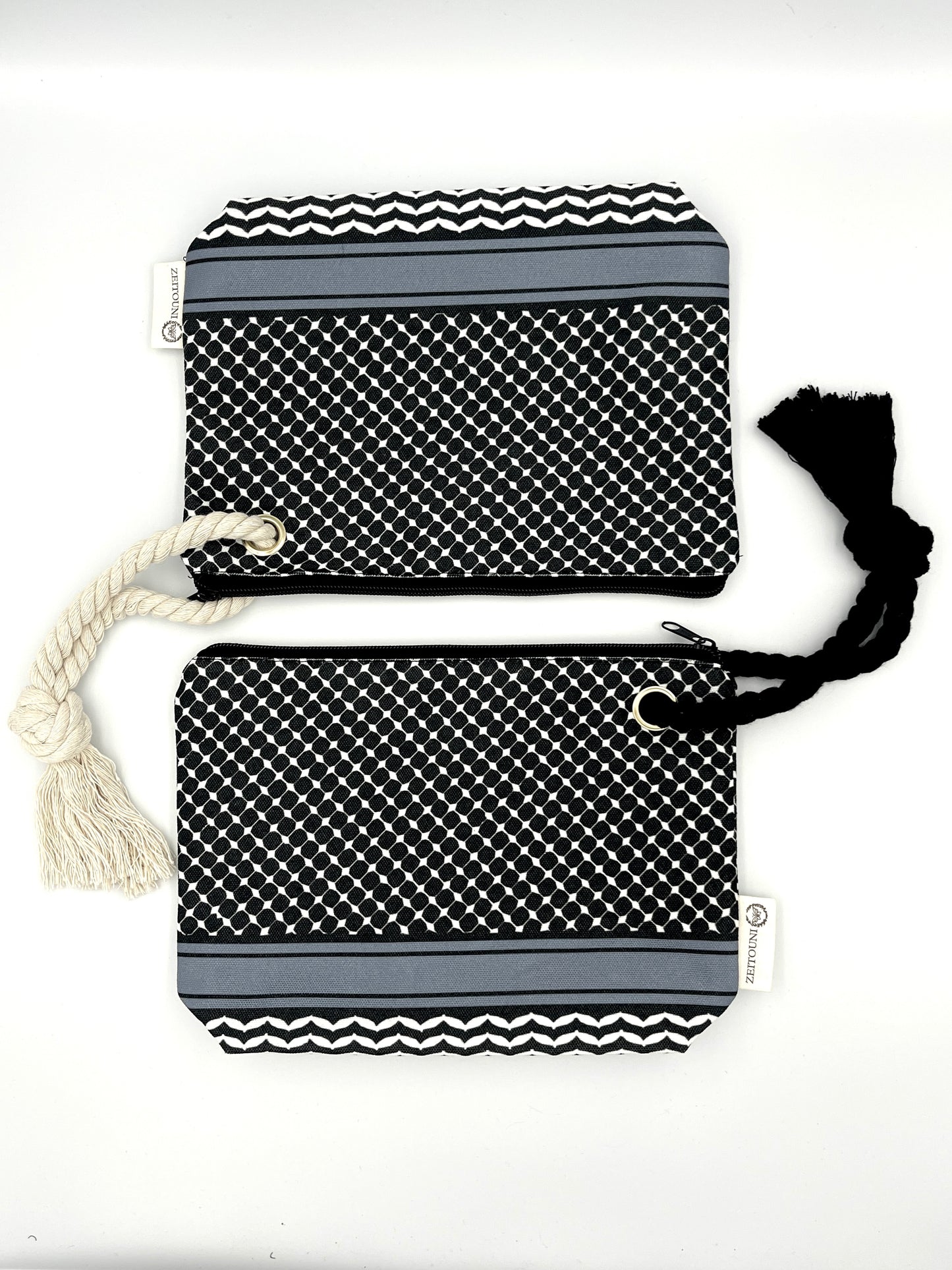 Two black makeup bags laying vertically to each other, with a black rope on the right attached to the bottom bag and a beige rope on the left attached to the top bag. Both bags are designed with a black keffiyeh pattern and ZEITOUNI logo tag. Laid against a white background.