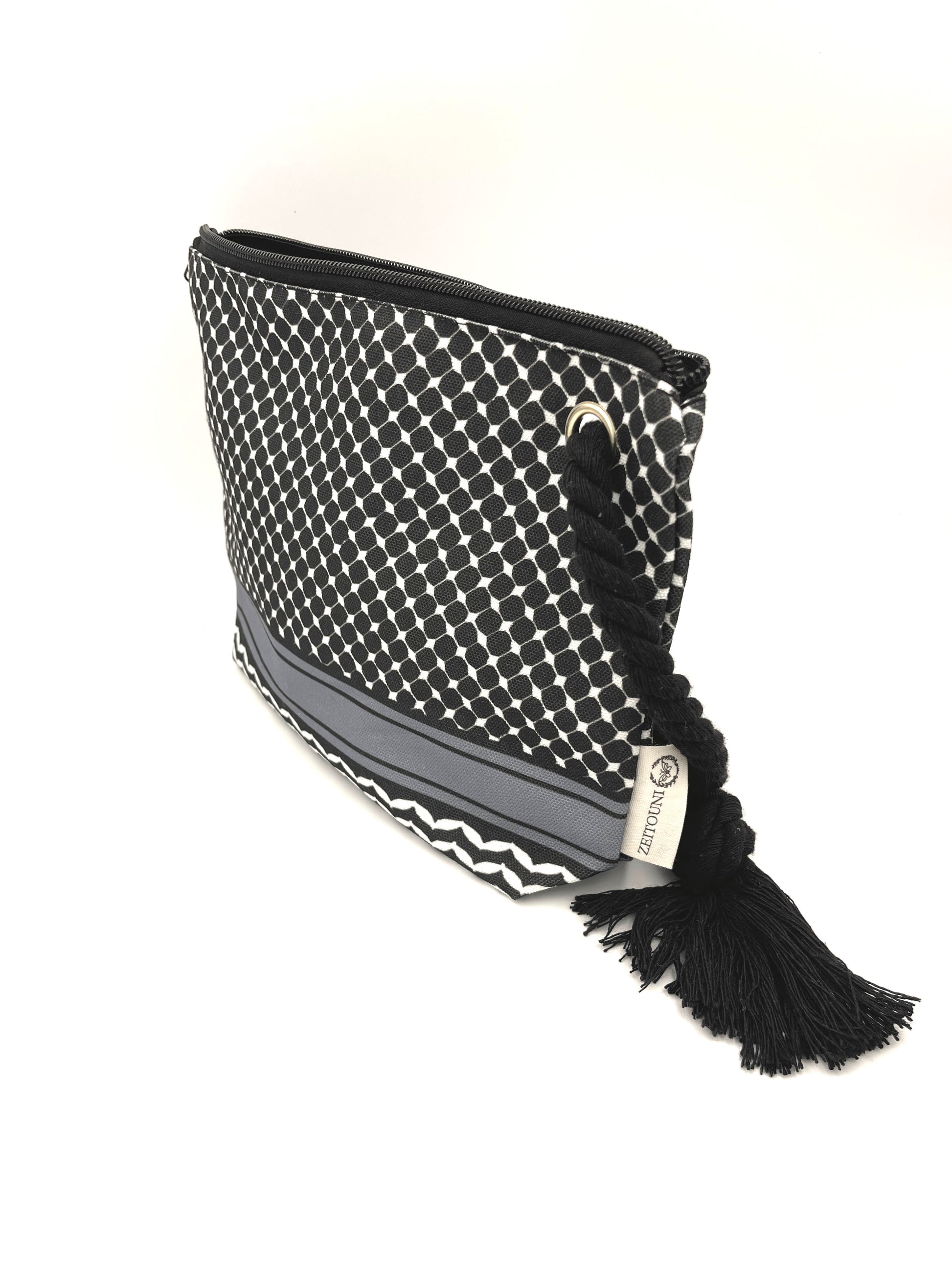 Angle shot of black makeup bag with black rope against a white background. Bag is decorated with keffiyeh pattern on pouch and ZEITOUNI logo tag.