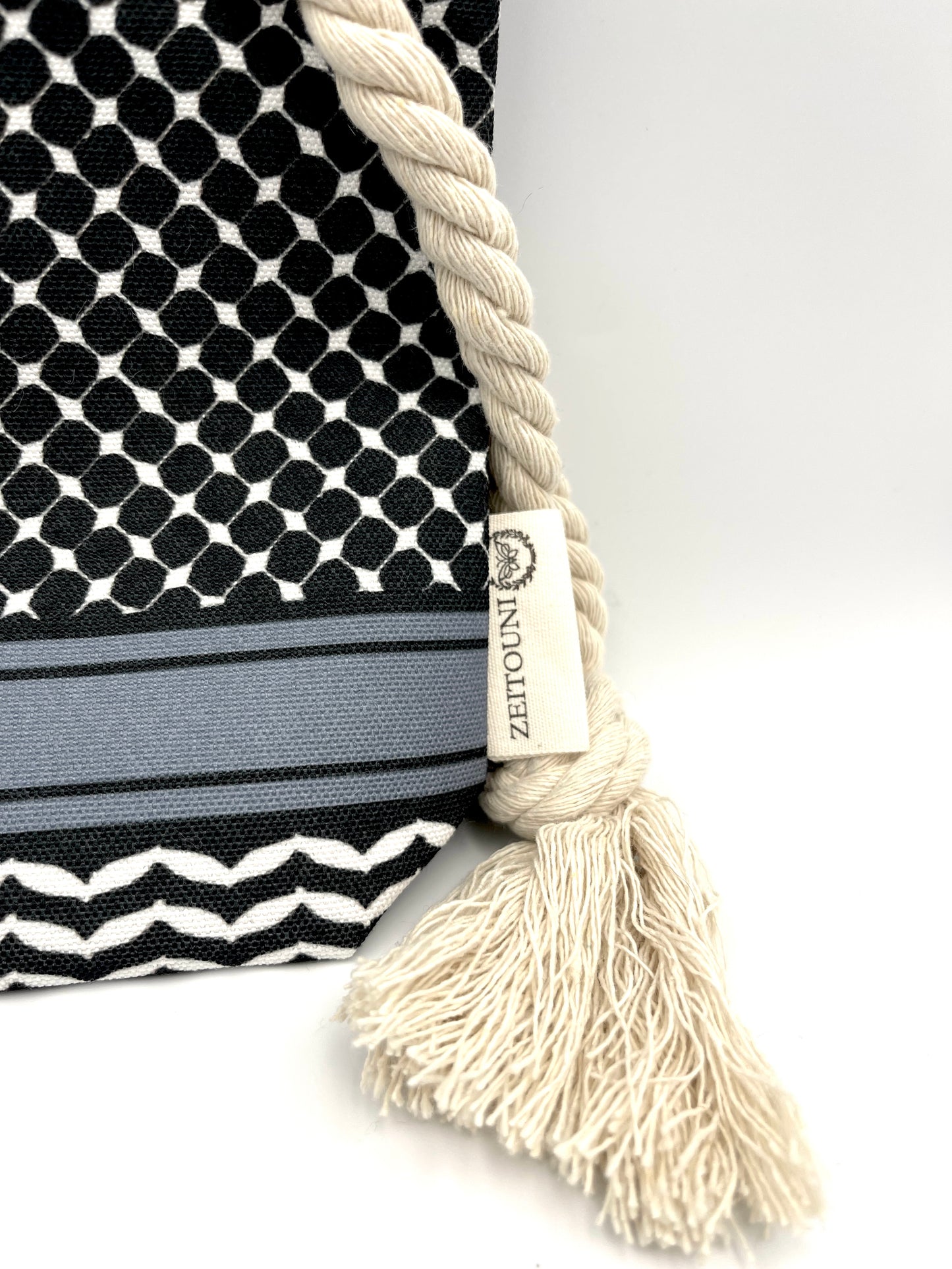 Macro shot of black makeup bag against white background showcasing rope, design, and tag. Bag is designed with a keffiyeh pattern on the pouch and a ZEITOUNI logo tag. 