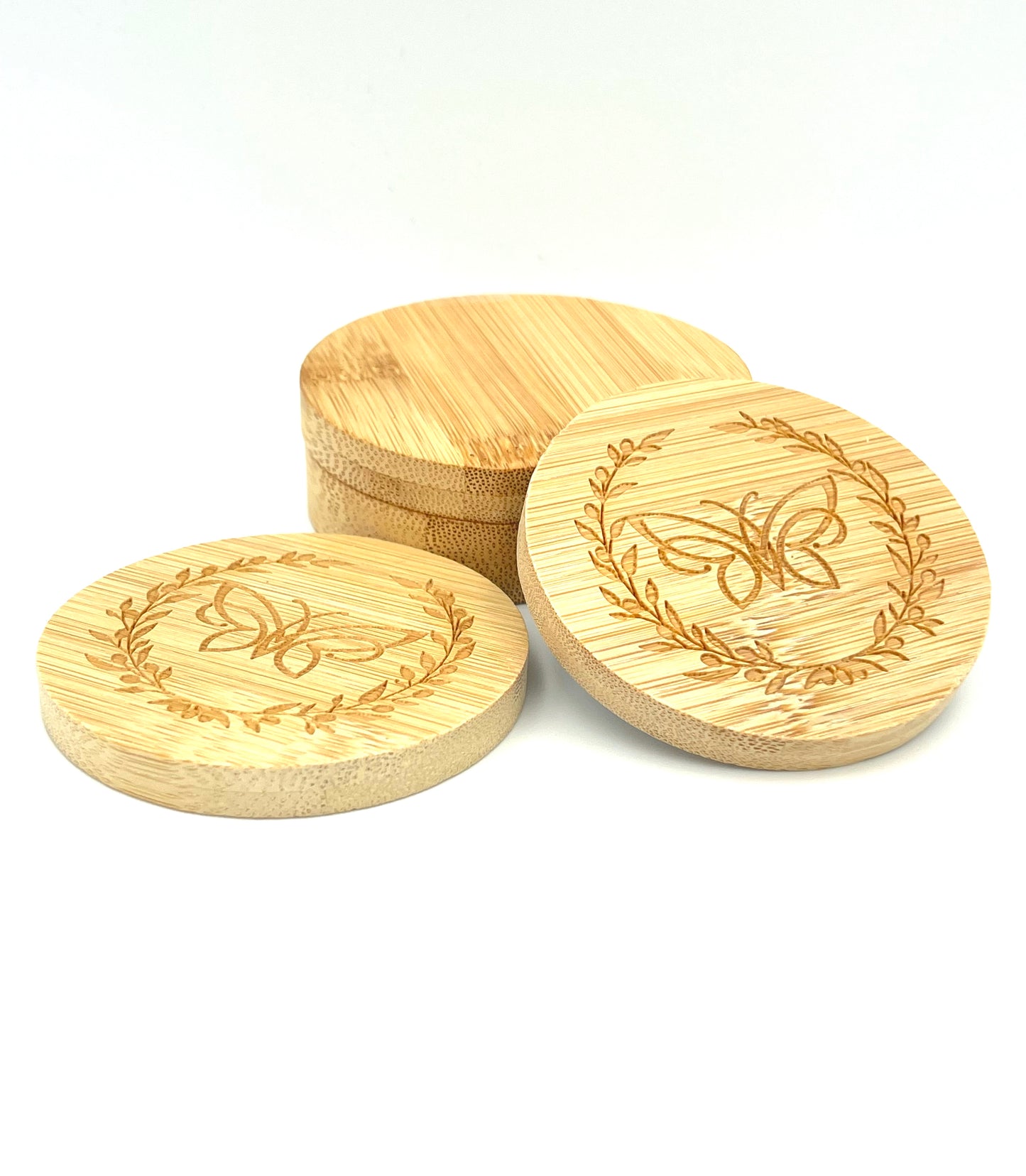 Two bamboo mirrors leaning against a bamboo case in front of a white background. Both mirrors are decorated with a logo comprising a butterfly surrounded by an olive wreath.