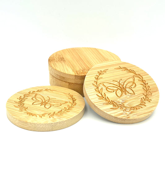 Two bamboo mirrors leaning against a bamboo case in front of a white background. Both mirrors are decorated with a logo comprising a butterfly surrounded by an olive wreath.