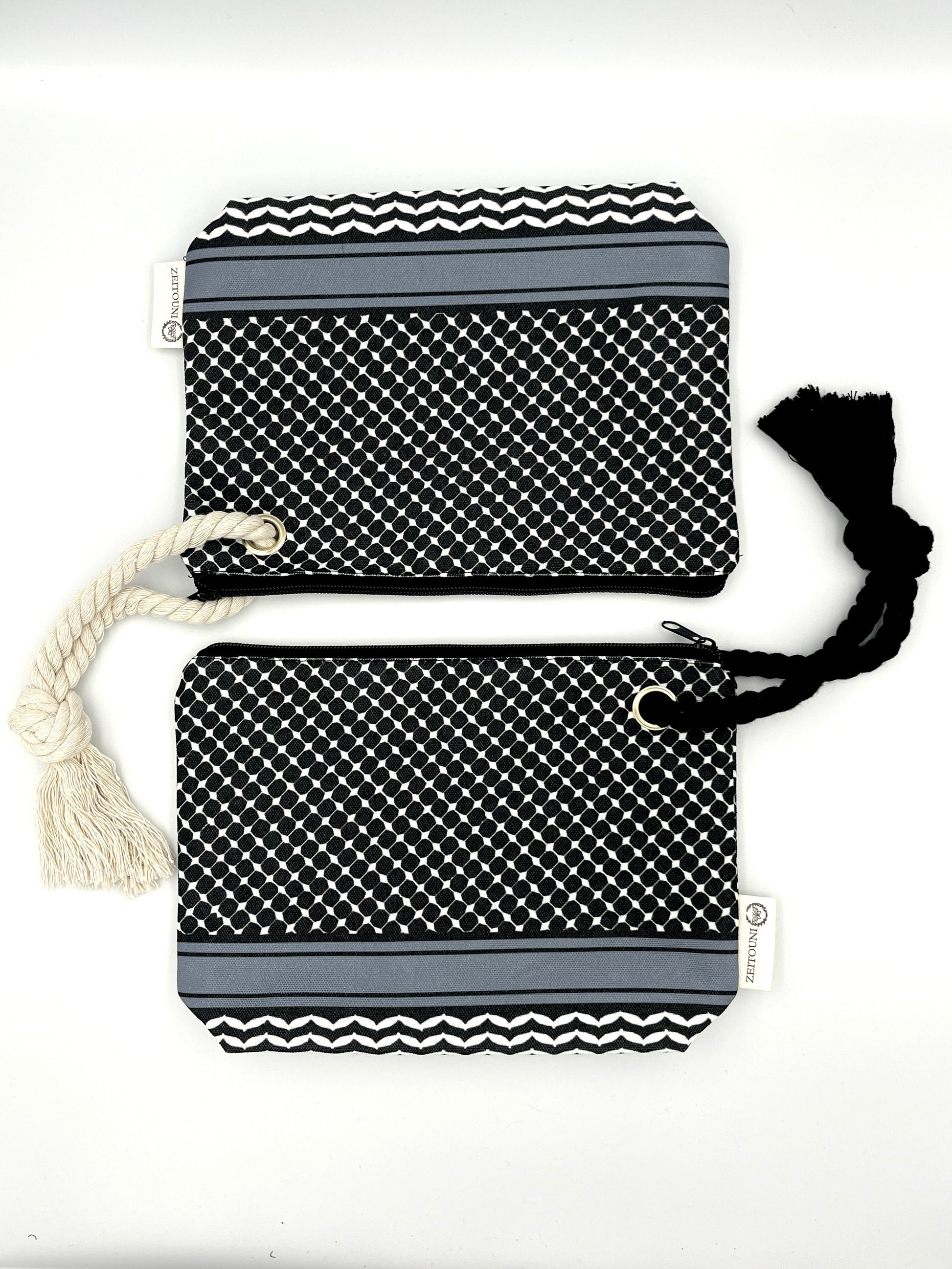 Two black makeup bags laying vertically to each other, with a black rope on the right attached to the bottom bag and a beige rope on the left attached to the top bag. Both bags are designed with a black keffiyeh pattern and ZEITOUNI logo tag. Laid against a white background. 