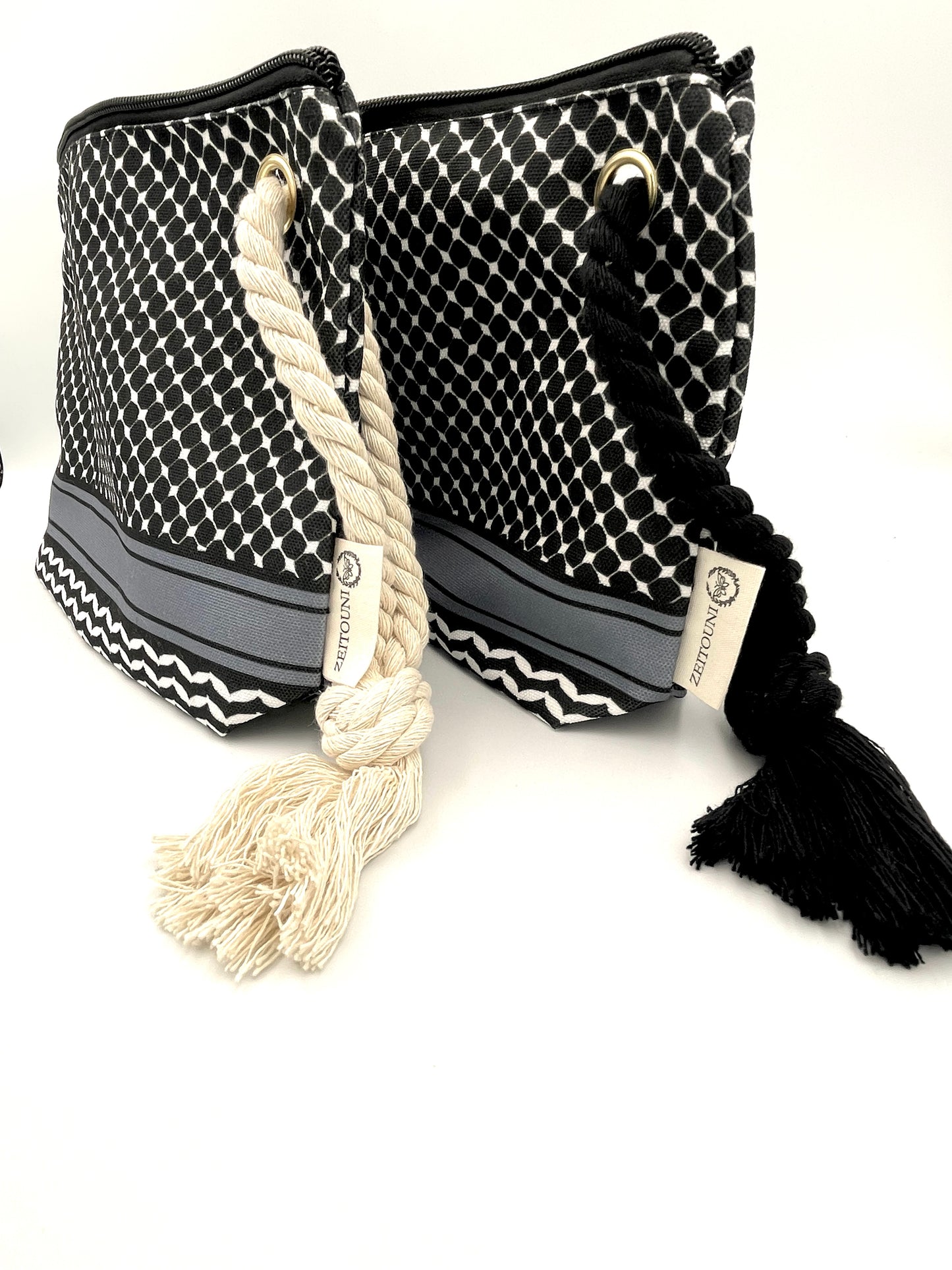 Angle shot of two black makeup bags against a white background. Left bag has beige rope, right bag has black rope. Both bags are designed with black keffiyeh pattern and ZEITOUNI logo tag. 