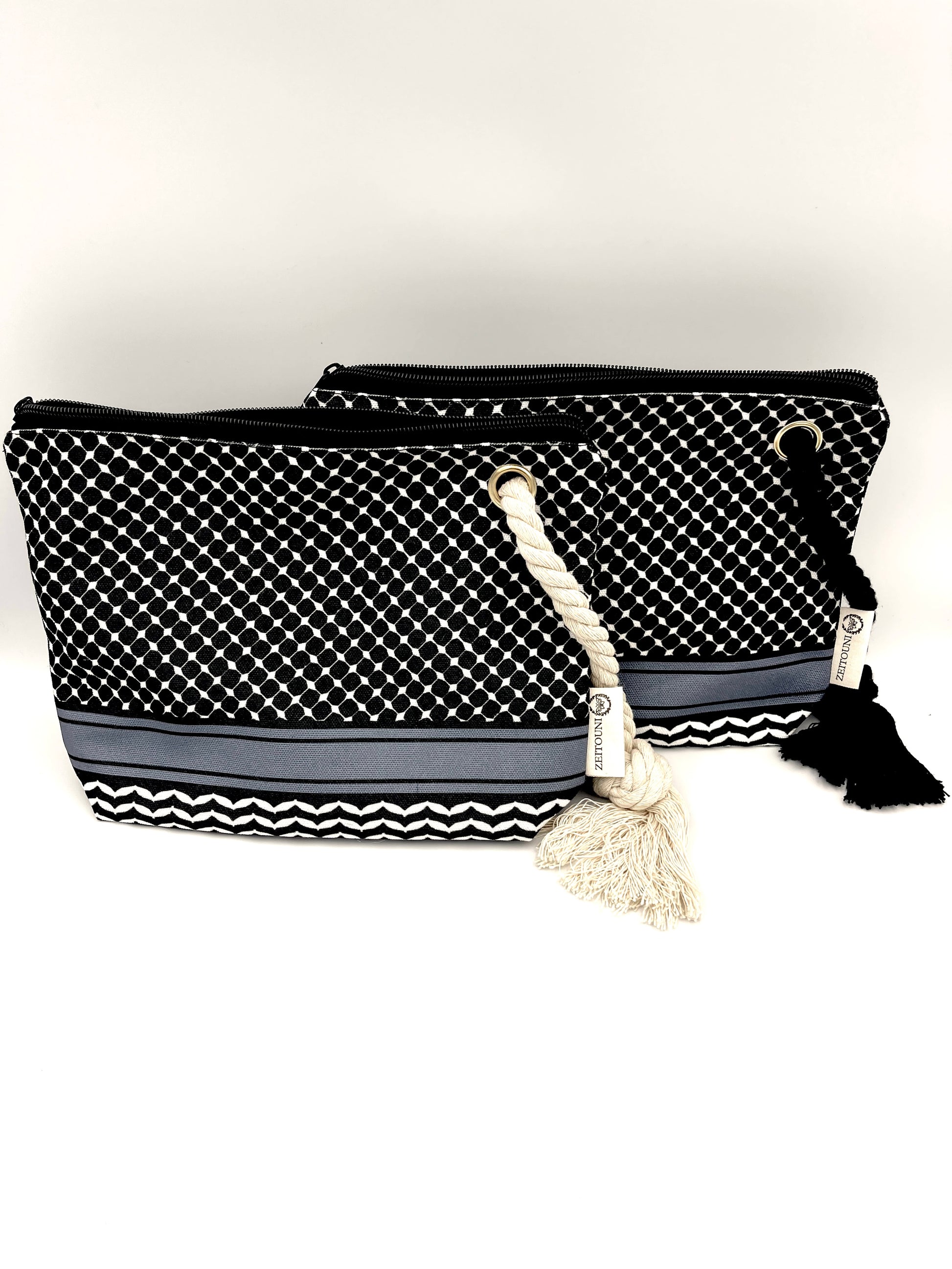 Front image of two black makeup bags laying in front of each other against a white background. Both are designed with a black keffiyeh background and ZEITOUNI logo tag. 